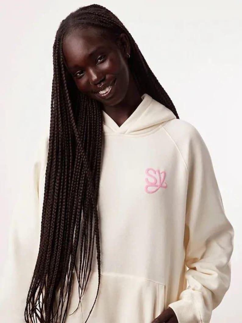 May include: Cream-colored hoodie with a front pocket and a hood. The hoodie features a pink embroidered logo on the chest. The model is wearing long, dark braids. The hoodie is made of a soft, comfortable material.