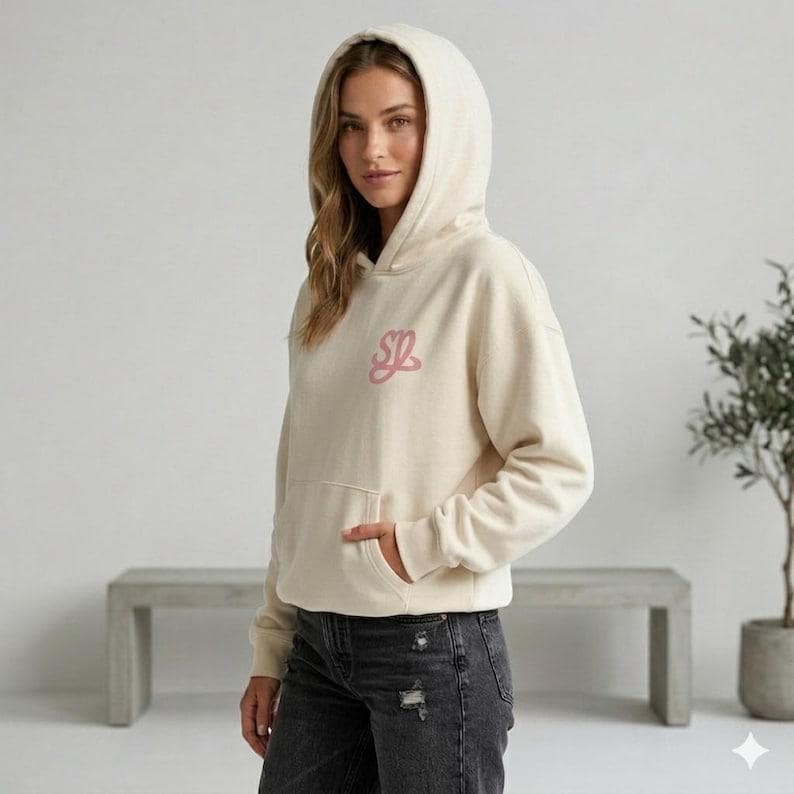 May include: Cream-colored hoodie with a front pocket and a hood. The hoodie features a pink embroidered design on the upper left chest. The person is wearing black jeans. The background includes a gray bench and a potted plant.