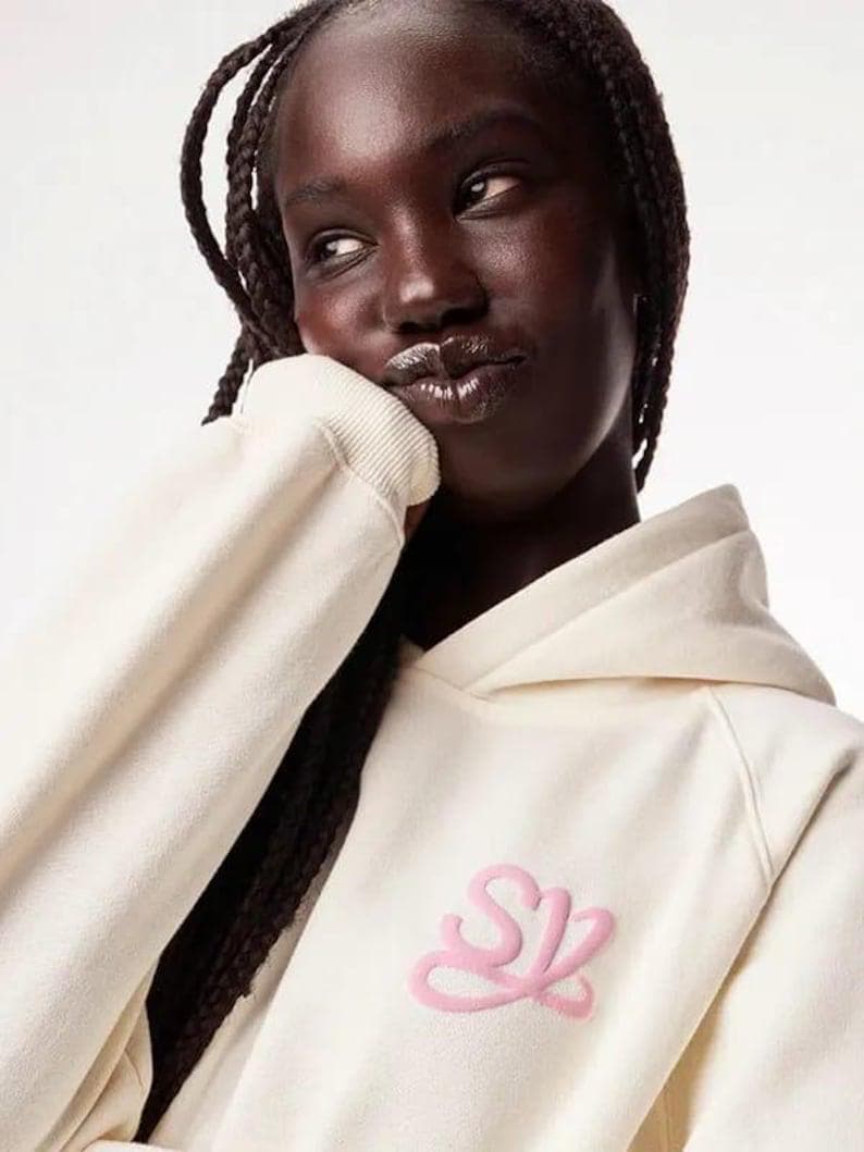 May include: Cream-colored hoodie with a pink embroidered logo on the front. The hoodie features a drawstring hood and long sleeves. The model is wearing the hoodie.