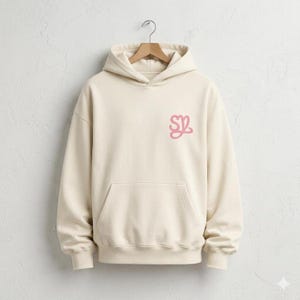 May include: Cream-colored hoodie with a front pocket and a drawstring hood. The hoodie features a pink stylized "SZ" logo on the left chest. The garment is hanging on a wooden hanger against a white wall.