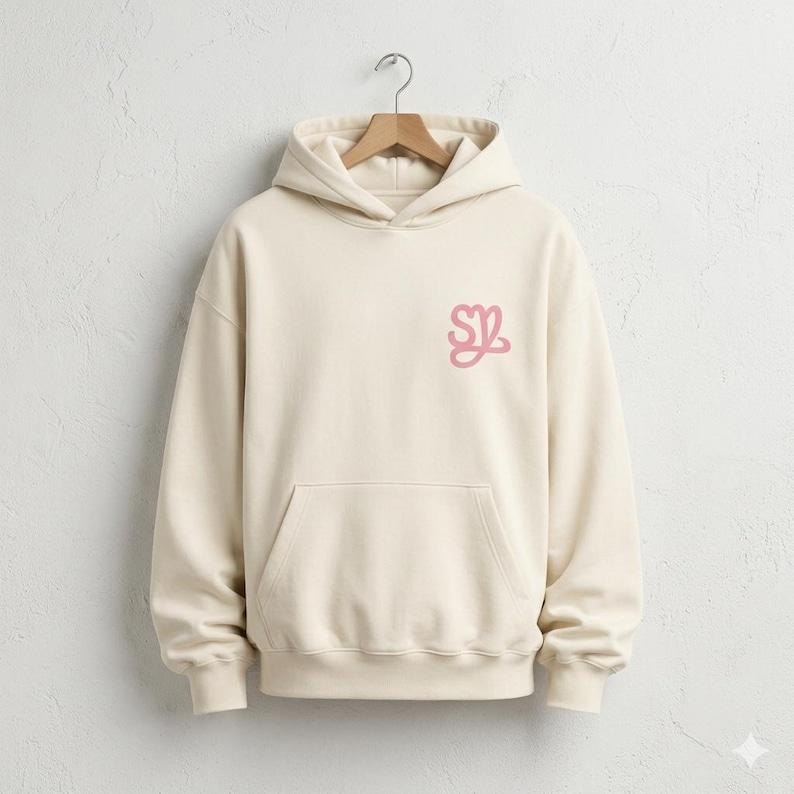 May include: Cream-colored hoodie with a front pocket and a drawstring hood. The hoodie features a pink stylized "SZ" logo on the left chest. The garment is hanging on a wooden hanger against a white wall.