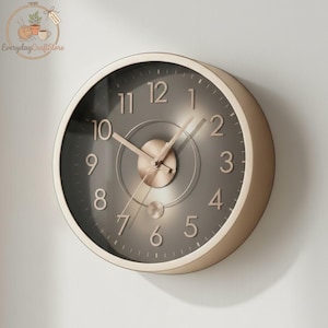 Puede incluir: Un reloj de pared redondo con un marco color crema y una esfera gris oscuro. El reloj presenta manecillas y números de color oro rosa. La esfera del reloj tiene un diseño circular en el centro. El reloj está montado en una pared blanca.