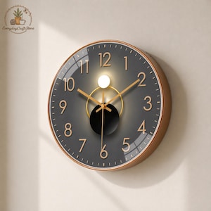 May include: Round wall clock with a grey face, gold numerals, and gold hands. The clock has a gold-coloured frame and a black centre circle. The clock is mounted on a light-coloured wall.