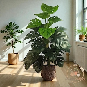May include: A large artificial Monstera plant with dark green, split leaves in a decorative brown pot. Another smaller plant in a tan pot is to the left. The scene is set in a well-lit room with wooden flooring.