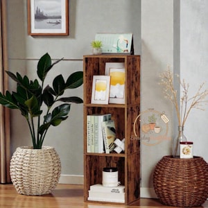 May include: A three-tiered, rustic brown wooden shelf unit. The shelves hold books, framed pictures, and a candle. A large potted plant in a woven basket sits to the left. A decorative basket and vase are on the right.