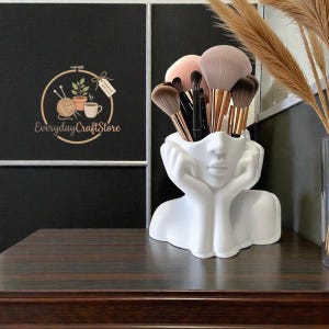 Op de afbeelding: Een witte, gezichtsvormige make-up kwastenhouder gevuld met diverse make-up kwasten. De houder staat op een donkerbruin houten oppervlak. De achtergrond toont een zwarte muur met een logo "Everyday Craft Store".