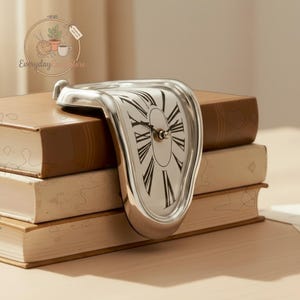 Puede incluir: Un reloj plateado y derretido con una esfera blanca y números romanos negros descansa sobre una pila de libros. El diseño del reloj es surrealista y artístico, con una apariencia distorsionada y fluida. Los libros son marrones y beige.