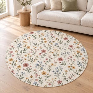 Può includere: Un tappeto rotondo con sfondo color crema caratterizzato da un motivo ripetuto di piccoli fiori di campo nelle tonalità del rosa, giallo e blu, accompagnati da steli e foglie verdi, che creano un disegno botanico sull'intera superficie.