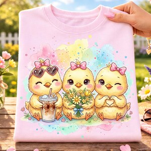May include: A light pink sweatshirt featuring a watercolor design of three cartoon chicks. One chick wears heart-shaped sunglasses and holds a drink, another holds flowers, and the third makes a heart shape with its hands.