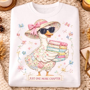 Może przedstawiać: Biała bluza z ilustracją kaczki w okularach przeciwsłonecznych i kapeluszu w kwiaty, trzymającej książki. Projekt zawiera kwiatowe detale i napis "JUST ONE MORE CHAPTER".