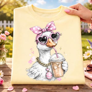 May include: A light yellow sweatshirt featuring a cartoon goose wearing heart-shaped sunglasses, a pink polka dot bow, and a gold chain. The goose is drinking from a clear cup with a pink straw. The design is whimsical and fun.
