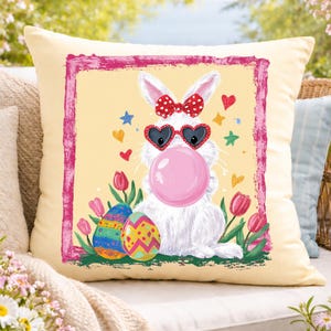 Może przedstawiać: Kwadratowa, kremowa poduszka dekoracyjna z kreskówkowym zajączkiem wielkanocnym dmuchającym bańkę z gumy do żucia. Zajączek nosi okulary przeciwsłoneczne w kształcie serca i czerwoną kokardę. Kolorowe pisanki i tulipany otaczają zajączka, z różową obwódką.