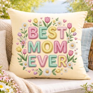 May include: A pale yellow square cushion featuring the text "BEST MOM EVER" in pastel shades, encircled by floral designs of tulips, daisies, and hearts. The cushion is placed on a white outdoor sofa.