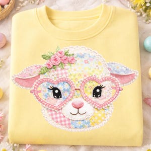 May include: A pale yellow sweatshirt with a patchwork sheep design. The sheep wears heart-shaped glasses and a floral crown. The patchwork includes floral and gingham patterns. The glasses are pink and white, creating a charming and playful aesthetic.