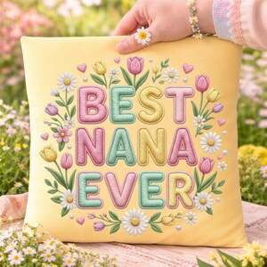 May include: A yellow square pillow with the words "BEST NANA EVER" in pastel colors, surrounded by floral illustrations of tulips, daisies, and leaves. The pillow is held up by a hand with a daisy ring and bracelet.