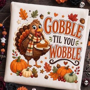 May include: A white fabric item featuring a cartoon turkey wearing a plaid scarf and holding a slice of pumpkin pie. The text reads GOBBLE TIL YOU WOBBLE in orange and brown letters, surrounded by illustrations of pumpkins, acorns, and autumn leaves.