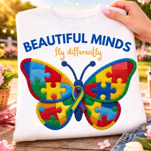 Może przedstawiać: Biała bluza z kolorowym motylem wykonanym z puzzli. Motyl ma niebieski tułów i czułki, ze skrzydłami z żółtych, czerwonych, zielonych i niebieskich puzzli. Tekst "BEAUTIFUL MINDS fly differently" jest wydrukowany nad motylem.
