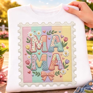 Puede incluir: Sudadera blanca con un diseño de patchwork colorido que deletrea "MAMA" dentro de un marco de sello postal. El diseño incluye patrones florales, corazones y un lazo, con un fondo a rayas pastel. La sudadera es sostenida por una persona.