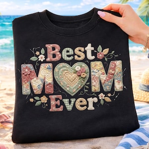 Peut inclure: Sweat-shirt noir avec l'inscription brodée "Best MOM Ever". Le texte est de style patchwork avec des motifs floraux et en forme de cœur. Le sweat-shirt est tenu par une personne sur une plage.