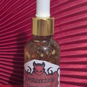 May include: A small glass bottle with a white dropper top and a gold-coloured cap. The bottle contains a liquid with visible particles and a label that reads "Destruction Hexing Oil" with a devil graphic. The background is a textured, deep red surface.
