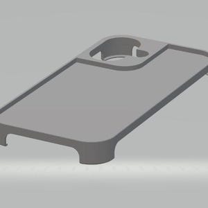 Puede incluir: Una funda de teléfono gris con un recorte circular incorporado y una bandeja rectangular. La funda tiene bordes redondeados y una superficie lisa. El diseño incluye dos ganchos curvos en el borde inferior.