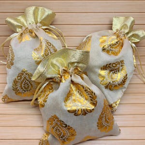 May include: Off-white fabric gift bags with gold paisley print and gold ribbon drawstring closures. Each bag measures 5.5 inches wide and 7.75 inches long. The bags are arranged on a light wood surface.