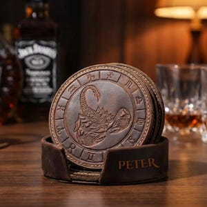 May include: A stack of brown leather coasters with a scorpion zodiac symbol embossed on each. The coasters are in a holder with the name "PETER" engraved. The coasters are round and have a decorative border with zodiac symbols.
