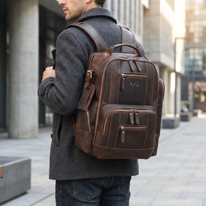 Op de afbeelding: Een bruine leren rugzak met meerdere vakken met ritssluiting en een handvat aan de bovenkant. De rugzak heeft een gepersonaliseerd monogram "A.G." op de voorkant. De tas wordt gedragen door een persoon in een grijze jas en een blauwe spijkerbroek.