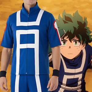 Op de afbeelding: Blauw jack met korte mouwen en bijpassende broek met witte accenten en een grote witte letter. De jas heeft een rits en rood-witte details op de mouwen. Een stripfiguur met groen haar staat op de achtergrond.