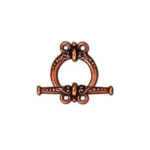 May include: A copper-colored toggle clasp with an ornate design. The clasp features a circular loop and a horizontal bar with decorative detailing. The metal has a warm, antique finish, suggesting a vintage or handcrafted aesthetic. The clasp is designed for jewelry or craft projects.