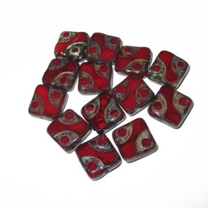 May include: A collection of square, red glass beads with a decorative silver and dark red design. The beads are arranged on a white surface, showcasing their glossy finish and intricate patterns. These beads are ideal for jewelry making.