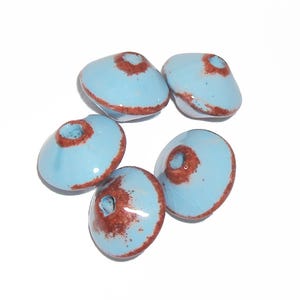 May include: Five light blue ceramic beads with a rusty brown accent around the central hole and edges. The beads have a smooth, glossy finish and are arranged against a white background. These beads are suitable for jewelry making.