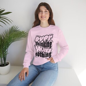 Puede incluir: Sudadera rosa claro con el texto negro "Overthinking Solves Nothing". La parte superior de manga larga la lleva una persona con vaqueros azules. La persona está sentada.