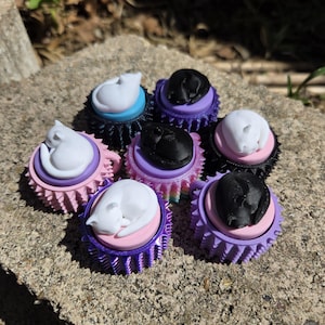May include: A collection of decorative cat-shaped toppers in various colors. Each topper features a sleeping cat figurine atop a circular base with a ruffled edge. Colors include white, black, pink, purple, and blue.
