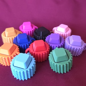May include: A collection of colorful, spiky-edged keycaps in various colors, including orange, blue, red, and green. Each keycap has a square top and a textured, gear-like base. The keycaps are arranged on a deep purple surface.
