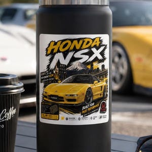 Może przedstawiać: Matowa czarna butelka termiczna z uchwytem na zakrętce, ozdobiona naklejką z żółtym samochodem Honda NSX, napisem Honda NSX oraz japońskimi znakami. Butelka stoi obok czarnego papierowego kubka do kawy, kluczyków samochodowych i okularów przeciwsłonecznych.