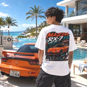 May include: A white short-sleeve t-shirt featuring a large graphic on the back of an orange Mazda RX-7 FD sports car. The design includes the text Mazda RX-7 FD, Rotary Rebellion, and Japanese characters, set against a dark, stylized background.