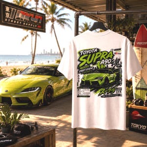 May include: A white short-sleeve t-shirt featuring a graphic print on the back. The design displays a lime green Toyota Supra A90 car with bold green and black text including 'Toyota Supra A90', 'Modern Street Legend', and Japanese characters.