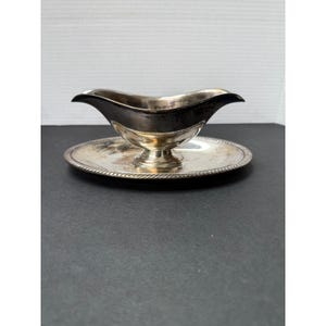 May include: A silver gravy boat with a black interior, resting on a matching round saucer. The boat has a curved handle and a pedestal base. The saucer has a decorative rope-like edge.