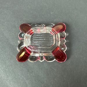 May include: A rectangular glass dish featuring a clear center with horizontal ridges and four rounded corners accented with deep red glass. The piece has a scalloped edge design and a smooth, reflective surface throughout its entire construction.