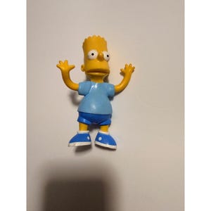 May include: A yellow plastic figurine of Bart Simpson with his arms raised. He wears a light blue t-shirt, dark blue shorts, and blue shoes with white accents. The figurine has a detailed face and spiky yellow hair.