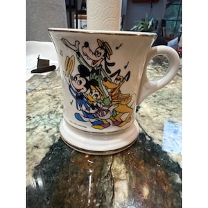 May include: Cream-colored ceramic mug featuring Mickey Mouse, Goofy, Donald Duck, and Pluto in a musical scene. The mug has a wide base, a curved handle, and gold trim. The words "Walt Disney Productions" are printed on the bottom.