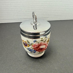 Pode incluir: Um porta-ovos vintage com um padrão floral. A chávena branca apresenta um desenho de flores vermelhas, azuis e roxas. Tem uma tampa prateada com uma pega. Um clássico dos utensílios de cozinha.