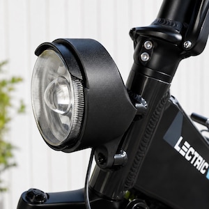 May include: Close-up of a black electric bicycle headlight. The round headlight has a clear lens and is mounted on the front of the bike. The bike frame is black, and the brand name "LECTRIC" is visible on the frame.