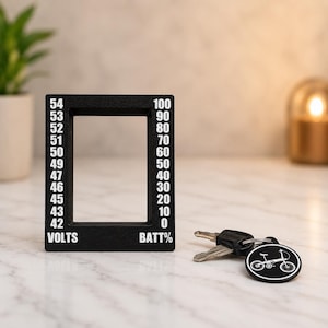 May include: A black frame with white text indicating voltage and battery percentage. The frame has a scale from 0 to 100 and 42 to 54. A set of keys with a bicycle-shaped keychain is next to the frame.