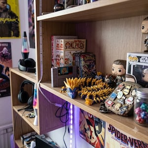 Könnte beinhalten: Ein Holzregal zeigt eine Sammlung von Gegenständen, darunter One Piece Manga-Bände, eine Super Nintendo-Spielkassette, eine gelb-blaue, bewegliche Drachenfigur, mehrere Funko Pop-Figuren, eine mit Anstecknadeln verzierte Tasche und ein Glas mit bunten Würfeln.