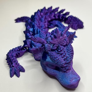 Könnte beinhalten: Eine 3D-gedruckte Drachenfigur in Lila- und Blautönen. Der Drache liegt, mit detaillierten Schuppen und Stacheln auf Rücken und Kopf. Die Figur hat ein strukturiertes Aussehen.