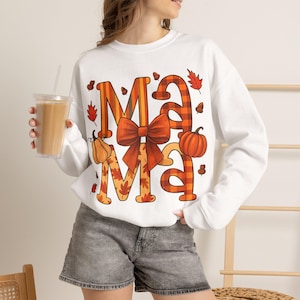 Könnte beinhalten: Ein weißes Langarm-Sweatshirt mit dem Wort MAMA in großen orange-braunen Karobuchstaben. Das Design zeigt eine auffällige rote Schleife in der Mitte, umgeben von orangefarbenen Kürbissen, braunen Eicheln und roten Herbstblättern.