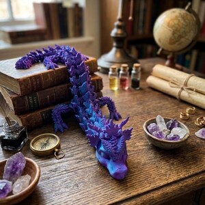 Könnte beinhalten: Eine flexible, bewegliche Drachenfigur mit einem lebendigen Farbverlauf aus violetten und blauen Schuppen. Der Drache ist auf einem Stapel alter Bücher positioniert, wobei sich sein langer, segmentierter Körper über den Holztisch erstreckt.