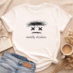 May include: Cream-coloured t-shirt with a black scribbled face design, featuring crossed-out eyes and a curved mouth. The text "mentally elsewhere" is printed below the face. The shirt is paired with blue jeans.